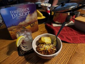 On a wooden surface, to the left of the frame, the game box for Forbidden Desert Thirst for Survival is standing. Up front if a town of chili with cornbread and a clear drink in a glass with butterflies on it, with a lemon slice. In the background is a pot and a pan filled with cornbread.