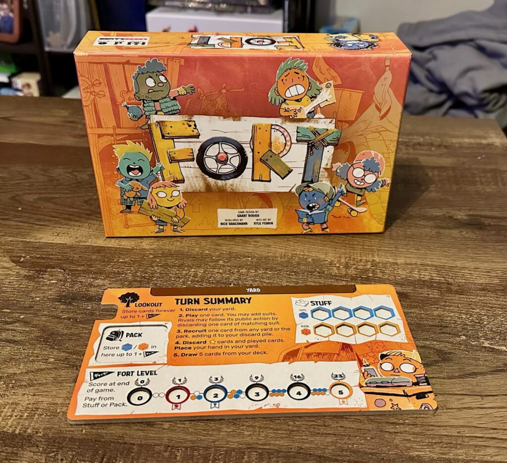 A tabletop display featuring the colorful Fort board‑game box in the background and a player reference card in the foreground. The card shows sections for Lookout, Pack, Stuff, Fort Level, and a detailed turn summary with icons and resource spaces.