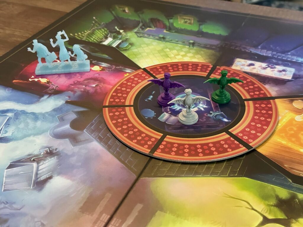 A circular hallway surrounds 3 bat style gargoyle meeples, colored purple, green and gray in the center. Rooms of different colors branch out from the center. A figure of 3 hitchhiking ghosts is off to the top left of the frame. 