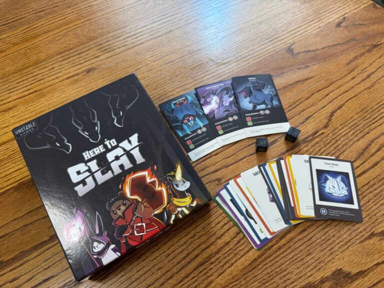 The front of the Game Boy for Here to Slay up Unstable Games is pictured, featuring a warrior bear, a mage bunny and a unicorn with a glowing horn. Next to the box the game cards and dice are spread out.