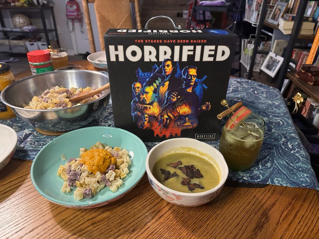 In the center of a table top is the Horrified board game. It reads The stakes have been raised on the top center of the box. Monsters such as Frankenstein and Dracula are shown on the center of the box. In the foreground is a cocktail, bowl of green soup and pasta with orange sauce. Off to the left side is a larger bowl of pasta. 