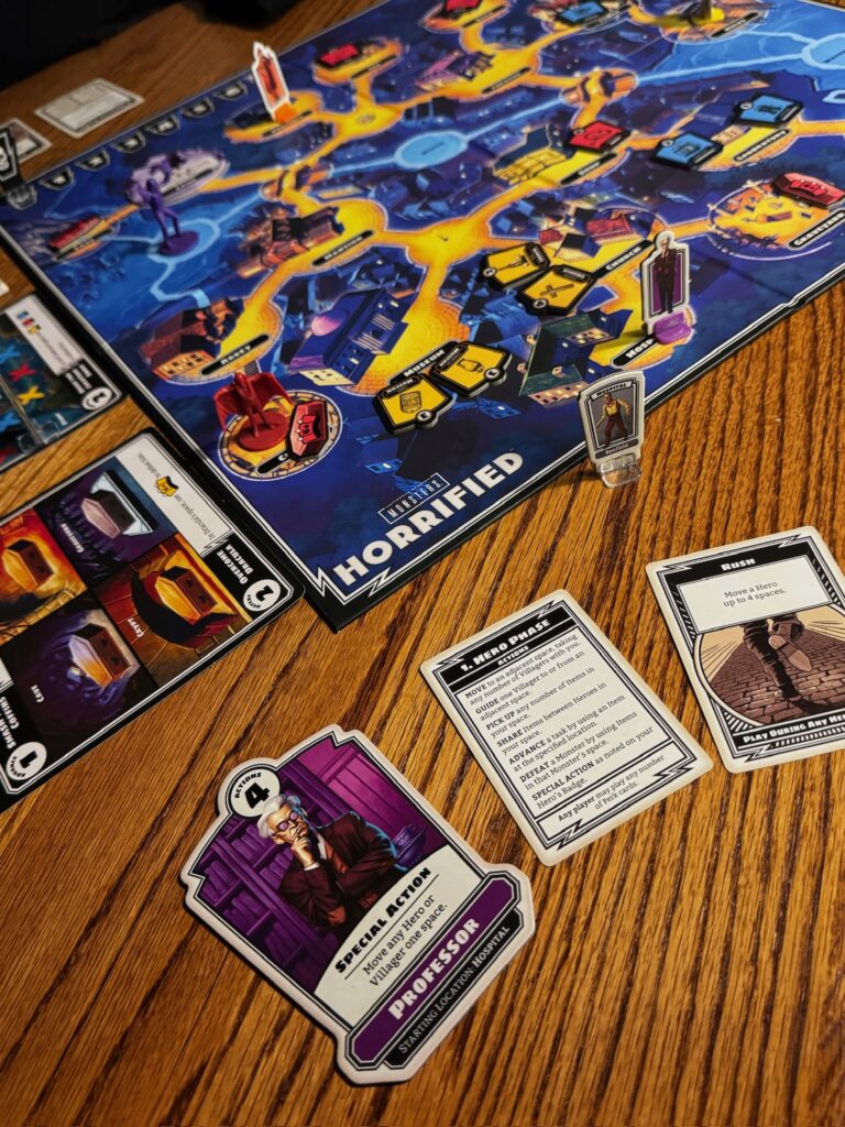 A game of Horrified is set up with a purple and red monster to the left and a purple and gray meeple to the right. Several cards are laid out around the board. 