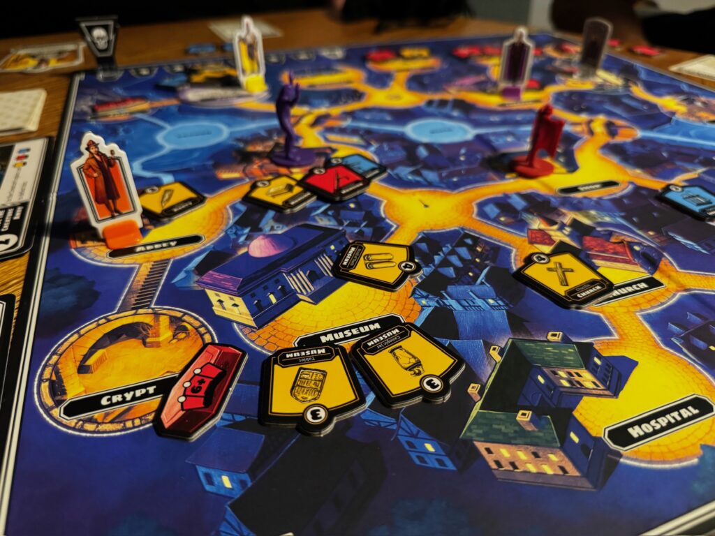 A close up of the Horrified game board depicts locations such as hospital, crypt and museum. Several meeples are around the board, indicating a game is in progress. 