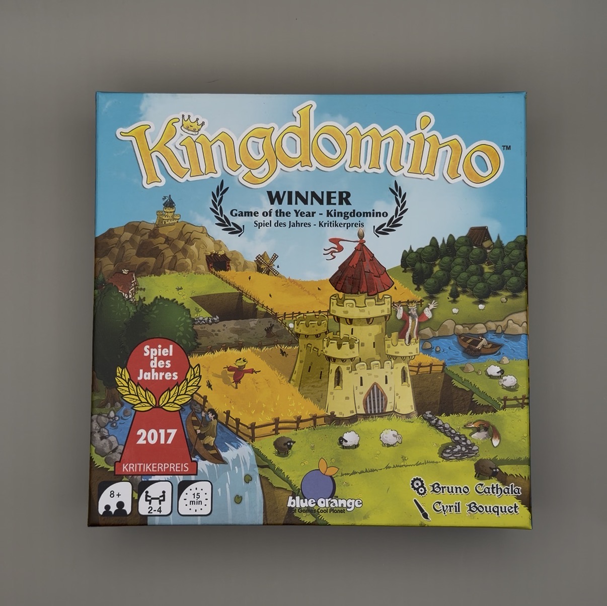 The box art of Kingdomino featuring Game of the Year 2017 Spiel des Jahres. The art features a castle in the middle with different patches of terrain surrounding it. A seal from blue orange games is in the bottom middle of the picture.