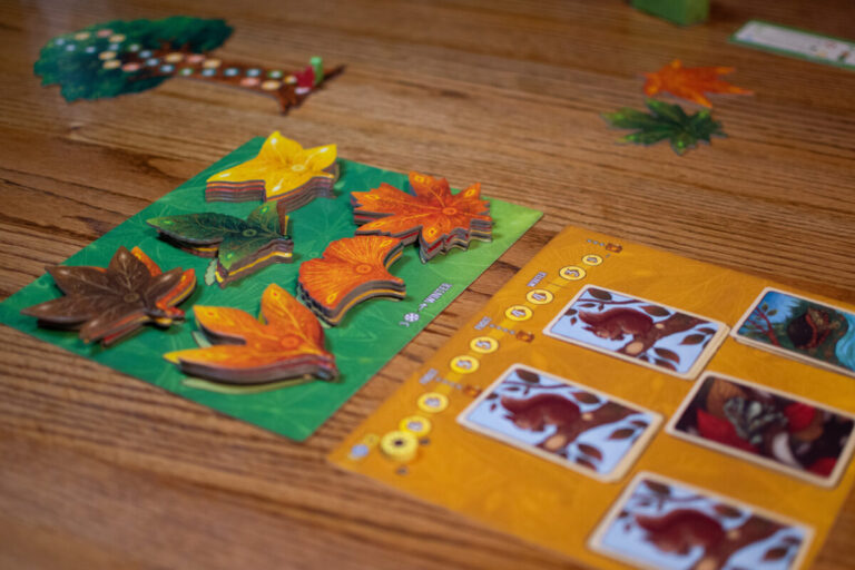 A close up of the game Leaf being played on a wooden table. One yellow card shows several forest critters on it, including three squirrels. A green card shows tiles of leaves in various shapes, in orange, yellow, green and brown colors. In the background is a tree score board.