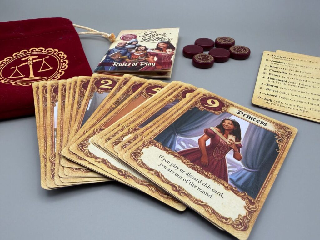 A close up of the game contents, focused on the character cards with the highest value card, the princess, sitting on top of the fan. The pouch, instructions, tokens, and reference cards are also shown behind the spread.