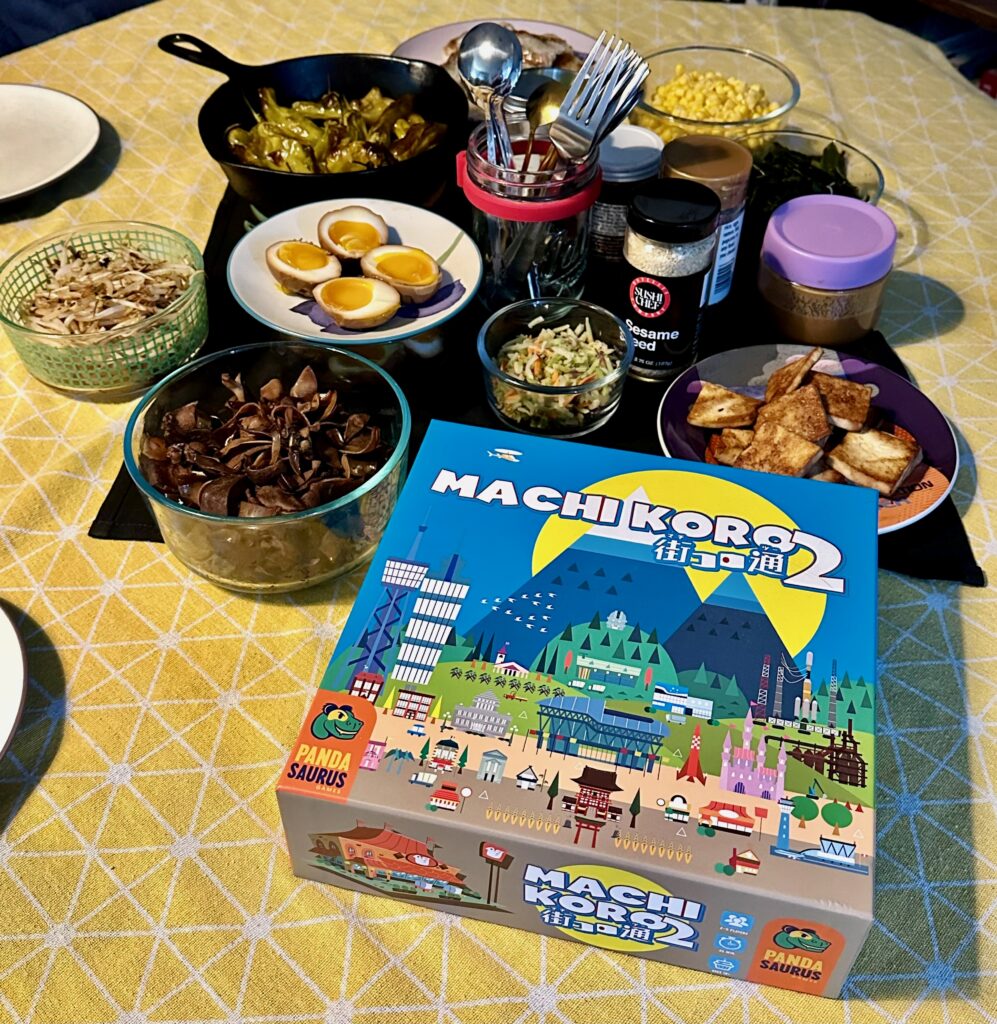 A themed dinner spread arranged on a dining table, featuring multiple bowls of prepared ingredients, soft-boiled eggs, roasted peppers, corn, herbs, sauces, and spring rolls, with the Machi Koro 2 board game box displayed in the foreground.