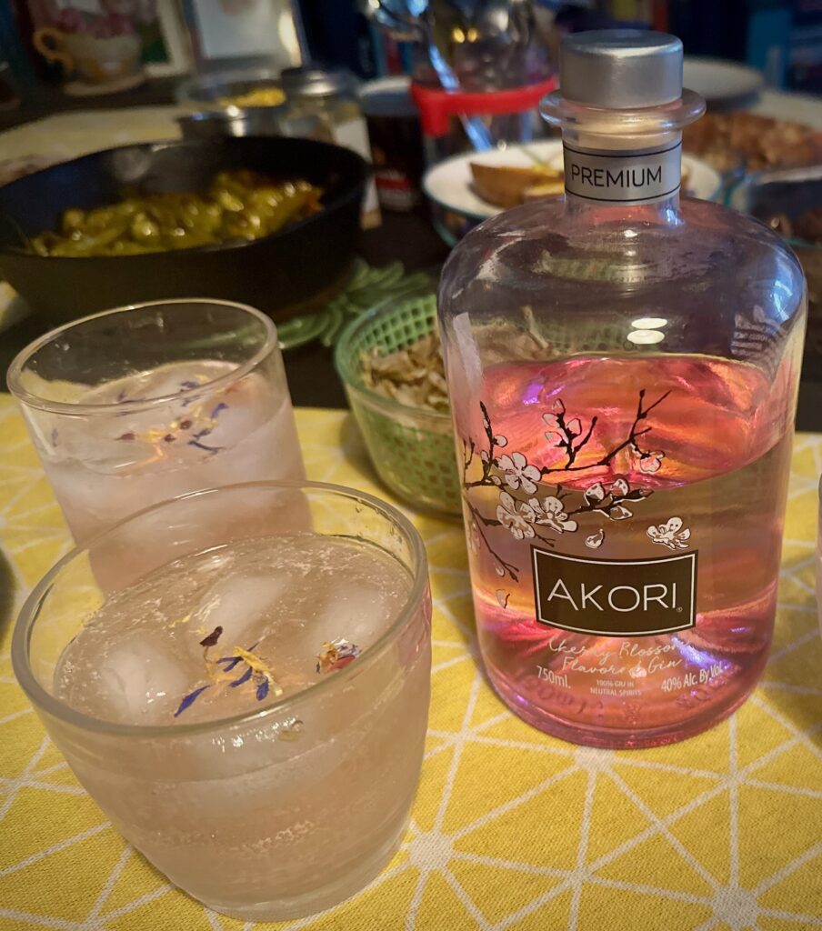 Two pale pink cocktails garnished with edible flowers sit beside a bottle of Akori Cherry Blossom flavored gin, arranged on a yellow patterned tablecloth during a themed game night dinner.