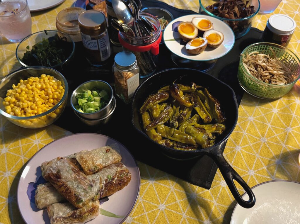 IMG_5361 A fully set dinner table featuring assorted bowls of vegetables, soft-boiled eggs, roasted peppers, corn, mushrooms, sauces, and spring rolls, arranged for a shared meal during a themed board game night.
