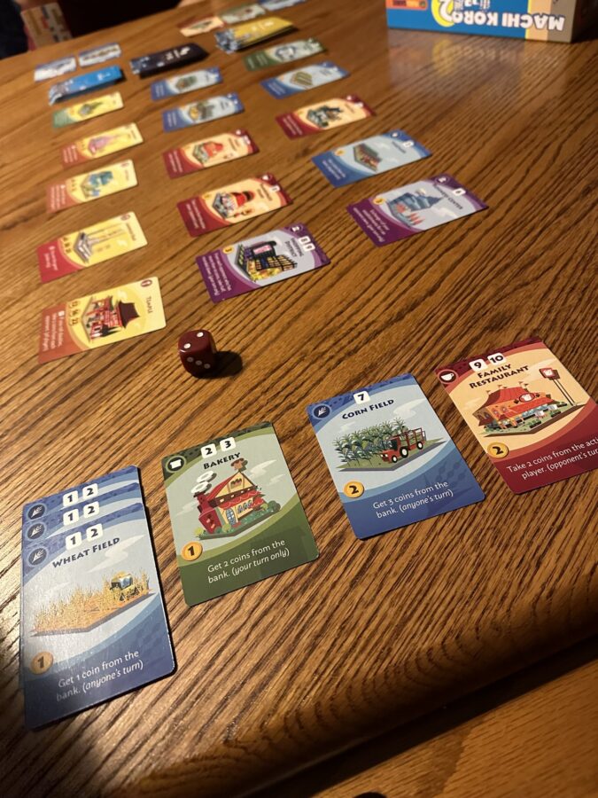 Cards laid out in rows across a wooden table during a game of Machi Koro 2, with player cards, establishment cards, and a single die visible, showing an in-progress game setup.