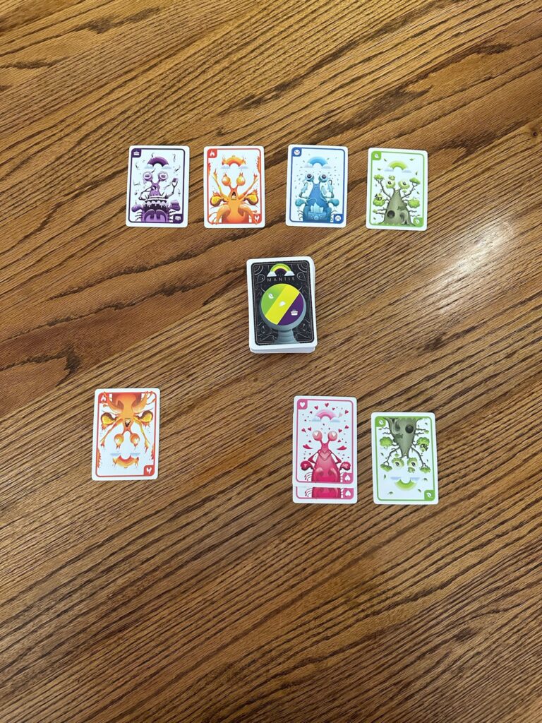 A set of colorful monster‑themed playing cards arranged on a wooden table, featuring creatures in green, blue, red, orange, and purple with numbers in the corners. A central deck sits in the middle with a top card showing a four‑color circular design.