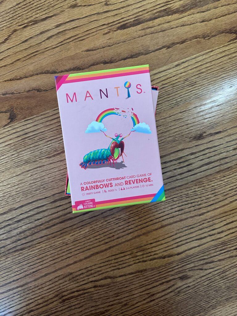 A colorful Mantis card‑game box resting on a wooden surface, featuring a stylized mantis surrounded by rainbows and clouds, along with text describing the game’s age range, player count, and publisher.