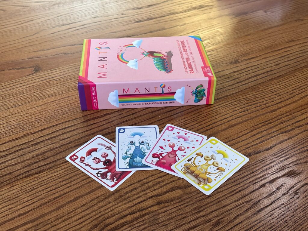 A Mantis card‑game box on a wooden surface with four brightly colored mantis character cards laid out in front. The cards show red, blue, pink, and yellow mantises, each with distinct themes such as skulls, water droplets, hearts, and confetti.