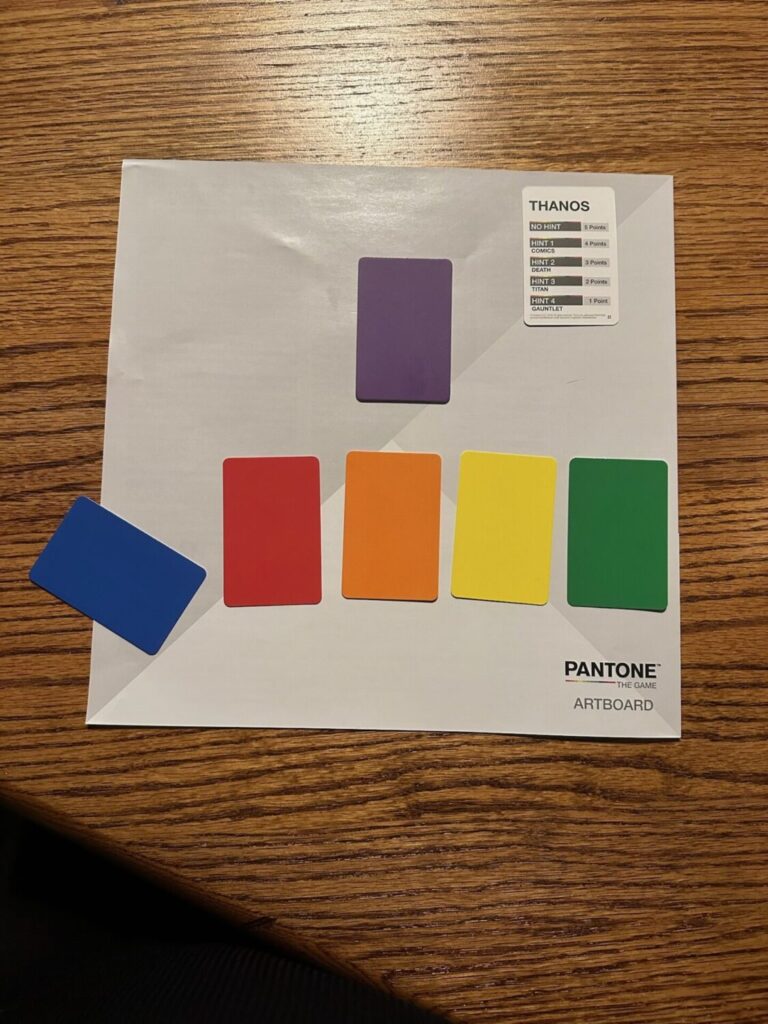 A card reading Thanos sits on top of a white playing area, on a wooden surface. Pictured are paint chips in purple, blue, red, orange, yellow and green. The bottom right hand corner of the white playing space reads "Pantone the game artboard."