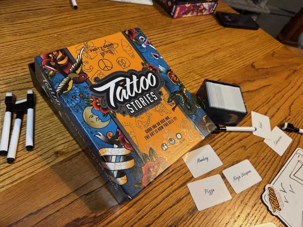 A game box labeled Tattoo Stories is pictured on a wooden table. The box reads good ink or bad ink... the art is how you sell it. Prompt cards reading monkey, pizza and ninja weapon are pictured along with a dry erase board and markers