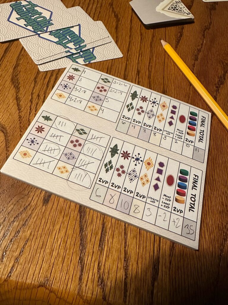 On top of a wooden table sits a pencil and a score pad with various designs on it. Off to the side are several playing cards that red That Old Wallpaper.