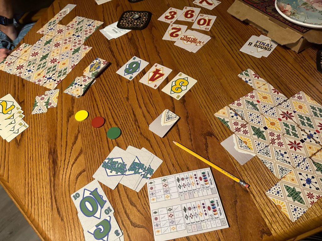 Thank Old Wallpaper is set up across a wooden table. Two diamond designs of tiles are at either side of the table with numbered cards reading 3, 4, and 6 in the center of the table. A Scorepad is also shown.