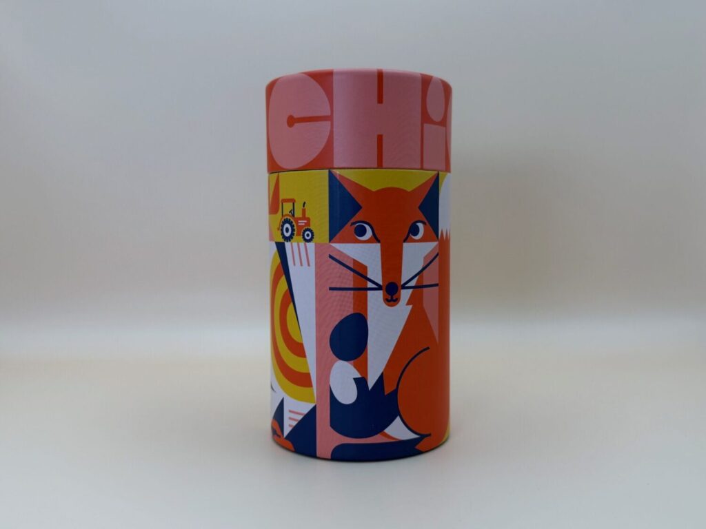 Photo of the chicken! game box, which is a cylinder with a vector-style fox holding two chicken eggs. The sides of the box are adorned with colorful patterns. The lid says "Chicken!"