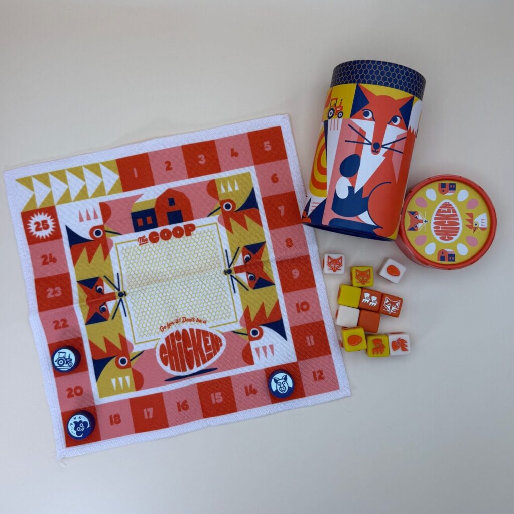 Game spread for Chicken! the game. It shows a cloth game board, several playing dice with different symbols, and the cylinder game box with the lid removed.