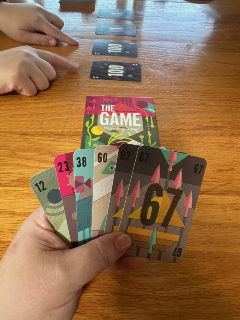 A tabletop game of The Game is in progress. A hand holds six numbered cards with colorful abstract designs, while four face‑up cards are laid out in a row on the table. Another player’s hand points toward the cards, and the game box sits nearby.