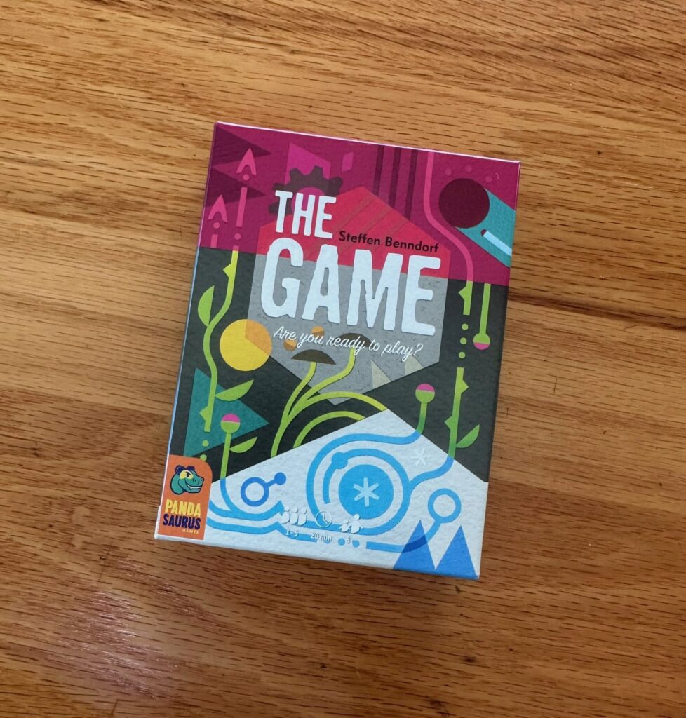 The board game box for The Game sits on a wooden surface, featuring bold, colorful abstract artwork with geometric and plant‑like shapes. The title appears prominently at the top, with player count, age range, and playtime icons along the bottom edge.