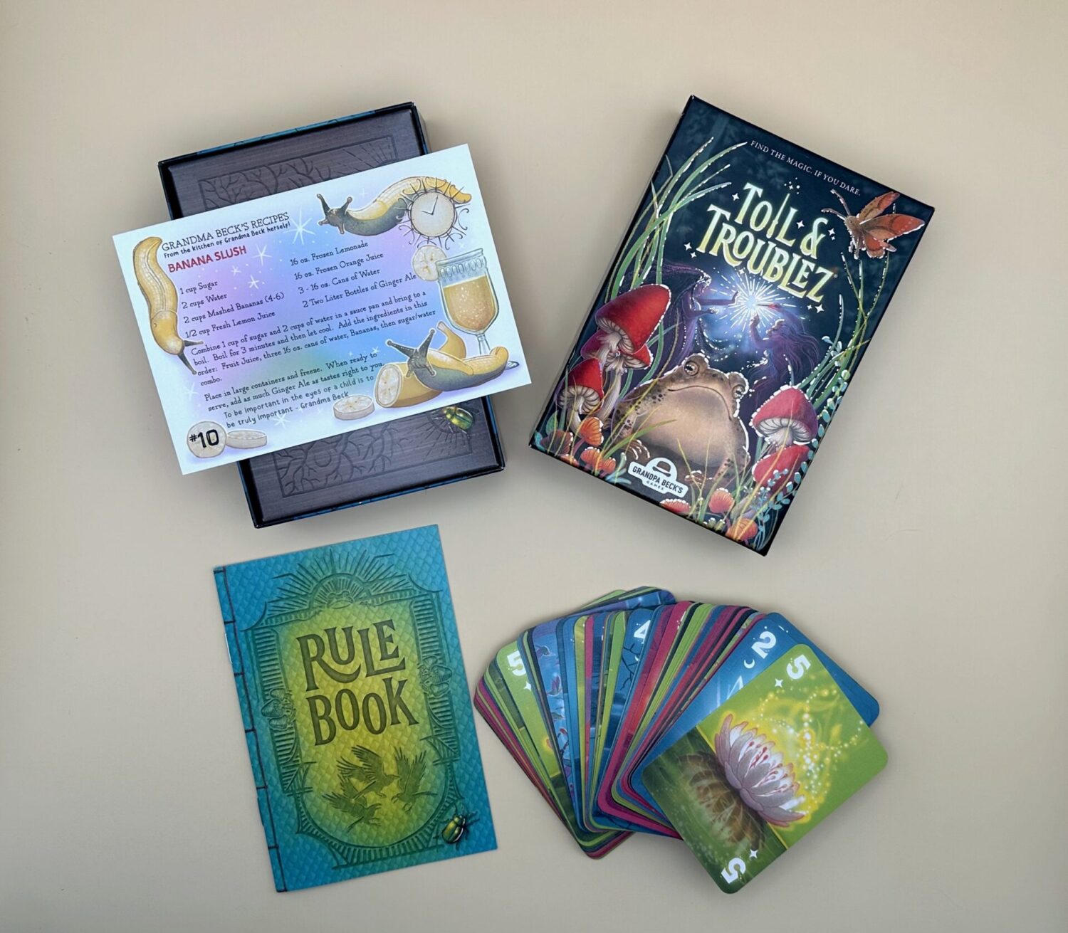 A close of up of the game box for Toil and Troubles is pictured. The artwork on the box features a large frog in between 2 growths of mushrooms. Also shown are a rule book, recipe for banana slush and game cards. The top card features a white Lilly opening and the number 5.