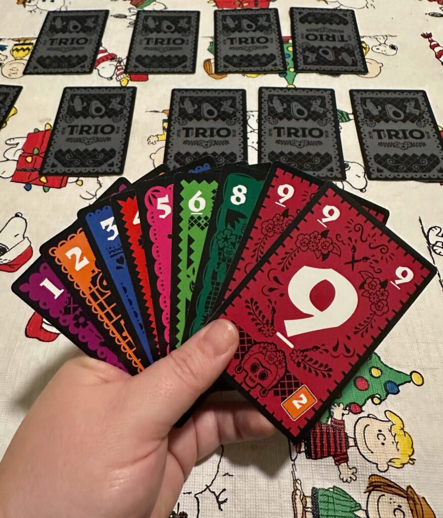 A hand is holding cards numbered 1, 2, 3, 4, 5, 6, 8, 9 and 9 in the foreground. In the background 9 Trio cards are spread out face down on a Snoopy and Friends table cloth. 