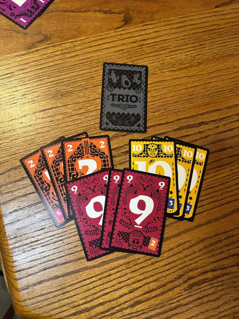 A Trio card is face down at the top of the frame. 3 sets of cards are shown: A Trio of 2, a Trio of 9 and a Trio 10. 