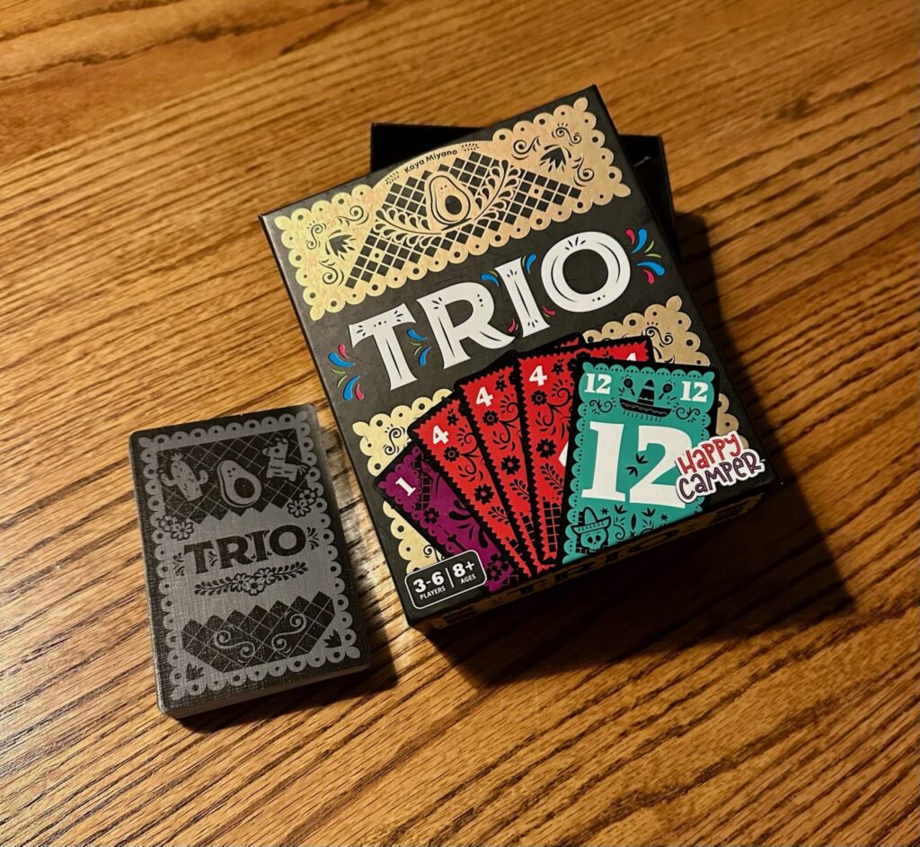 The center of the picture shows the Trio game box, with cards numbers 1, 4, 4, 4, and 12 showing. The Happy Camper Logo is shown at the bottom right and an icon showing 306 players and ages 8+ is on the lower left hand side of the box. To the left of the game box is the back of one of the Trio cards with Trio in the middle and a cactus, avocado and piñata decorating the top of the card. 