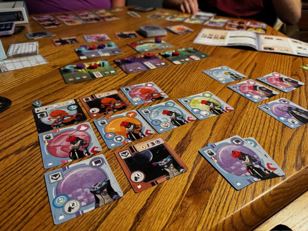 The Wild Space game is set up on a wooden surface. Many character cards are drafted on the table. In the background a rule book and scoring pad are also present.