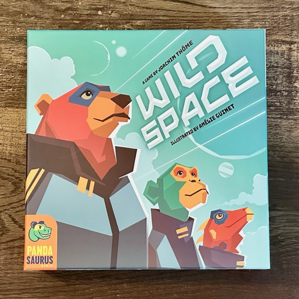 The board game cover for Wild Space is pictured on a wooden surface. The Pandasaurus logo is on the bottom left corner of the box. A bear, monkey and dragon dressed as astronauts are on the front of the box with a blue background of varying shades.