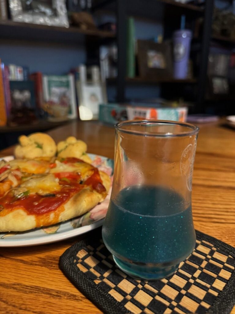A glittery, teal drink is pictured in a glass, on top of a coaster. Peeking on the left side of the frame is pizza on a floral plate.
