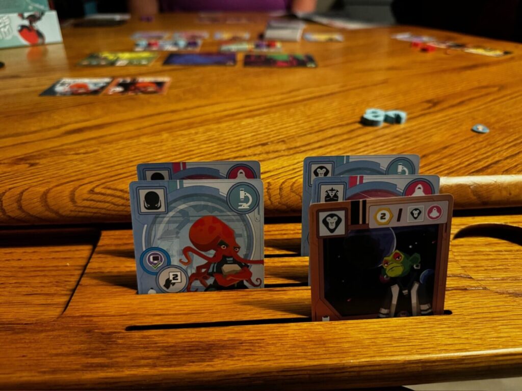 On a wooden game table, wild space cards are present. In the foreground creature cards of an octopus and monkey dressed as astronauts are pictured. In the background, various cards are on the playing area.