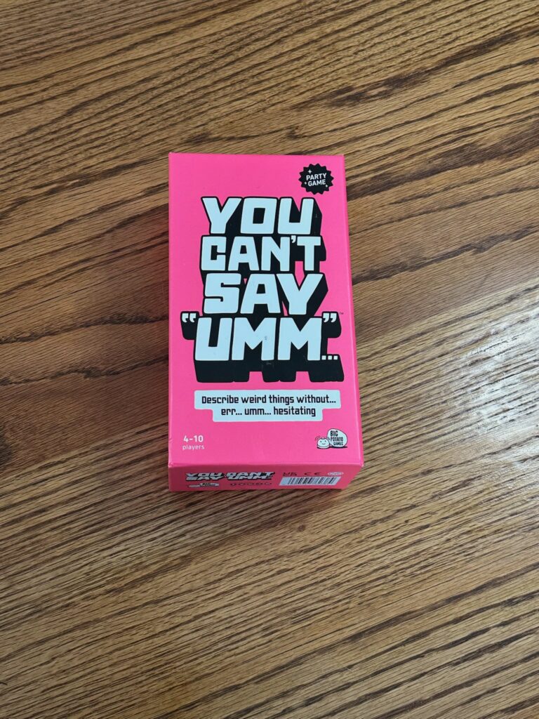 A bright pink “You Can’t Say ‘Umm’” party‑game box on a wooden surface, featuring bold black and white text and a starburst icon indicating it’s a party game.