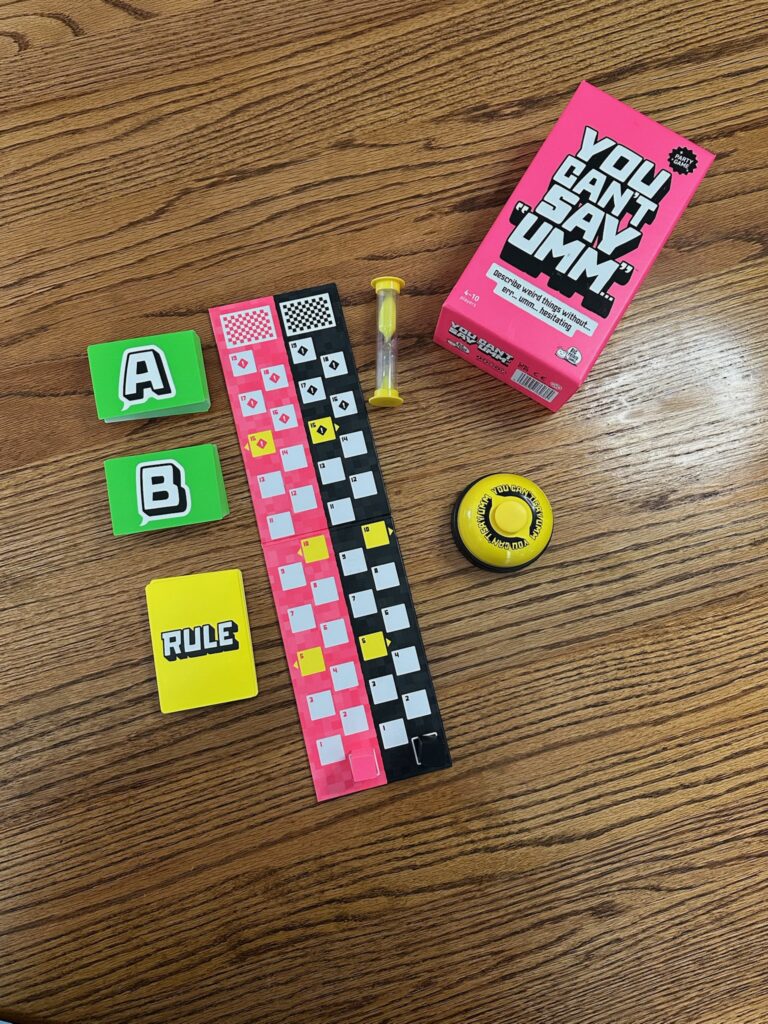 A party game setup displayed on a wooden table, including a bright pink game box, a yellow sand timer, a yellow buzzer, a colorful game board with question‑mark spaces, and three small decks of cards labeled A, B, and Rule.