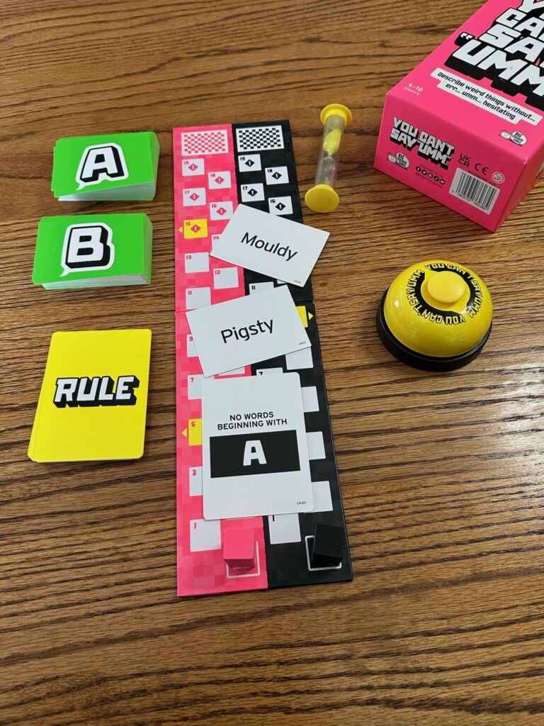 A tabletop display of the “You Can’t Say Umm” party game, including a bright pink game box, a yellow buzzer, a yellow sand timer, a game board with numbered spaces, colored player markers, decks of cards labeled A, B, and Rule, and several word cards laid out such as “Mouldy,” “Pigsty,” and a rule card reading “No words beginning with A.”