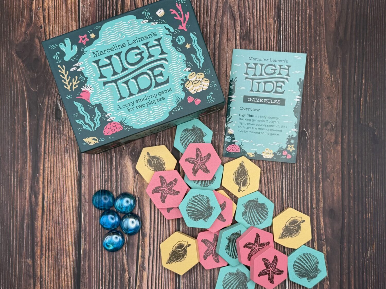 The box for the game High Tide in the the back of the frame. A rule book is pictured. In front of the box are yellow, blue and pick tiles, all with pictures of sea shells and starfish. 5 glass stones are on the left of the tiles.