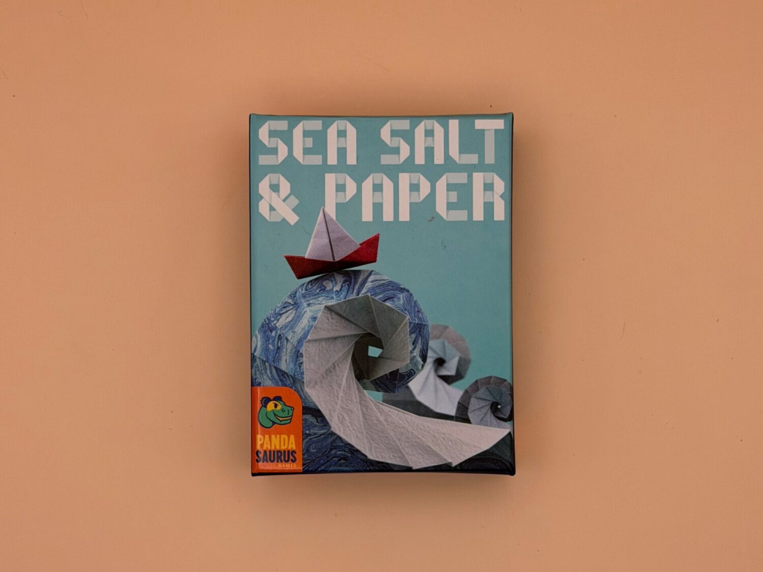 Pictured is the game box for Sea Salt and Paper with the Pandasaurus logo on it. On the box is an origami sailboard on top of origami waves.
