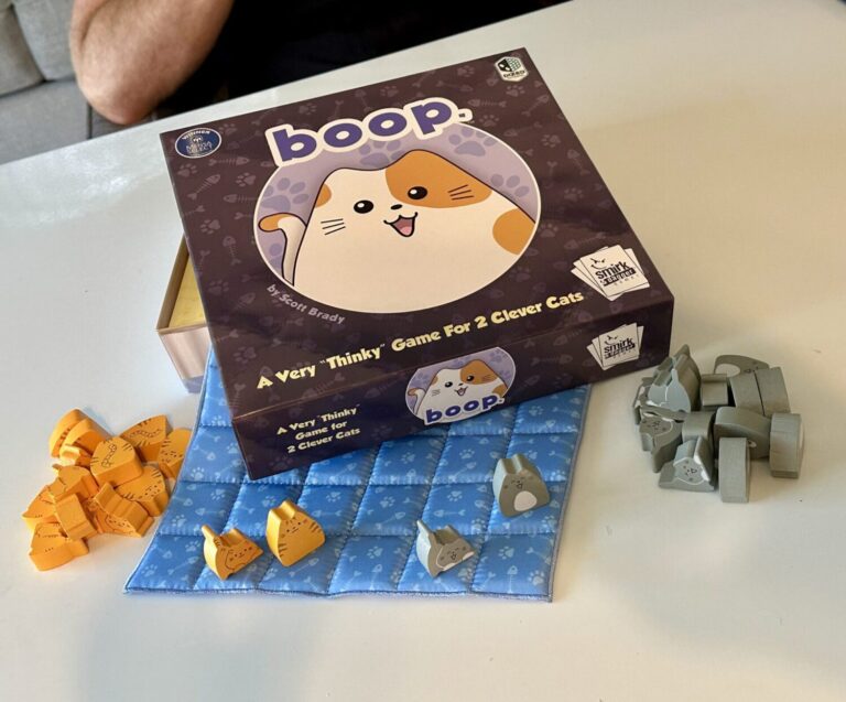 The boop game box is open and two piles of cats and kittens, one gray and one orange, sit aside the box. Several of the cat and kitten figures are on a blue quilt that is peeking out from under the box. The box features a cartoon calico cat and the phrase "a very thinly game for 2 clever cats."