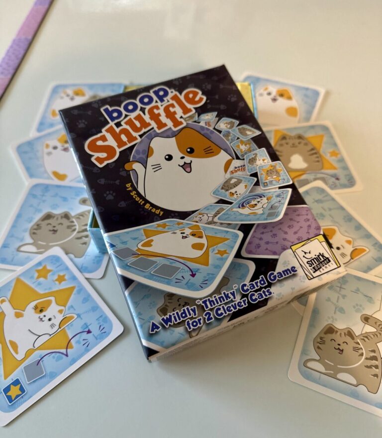 The boop shuffle game box sit onto of a pile of boop shuffle playing cards. The box features a cartoon calico cat and the phrase "a wildly thinky card game for 2 clever cats."