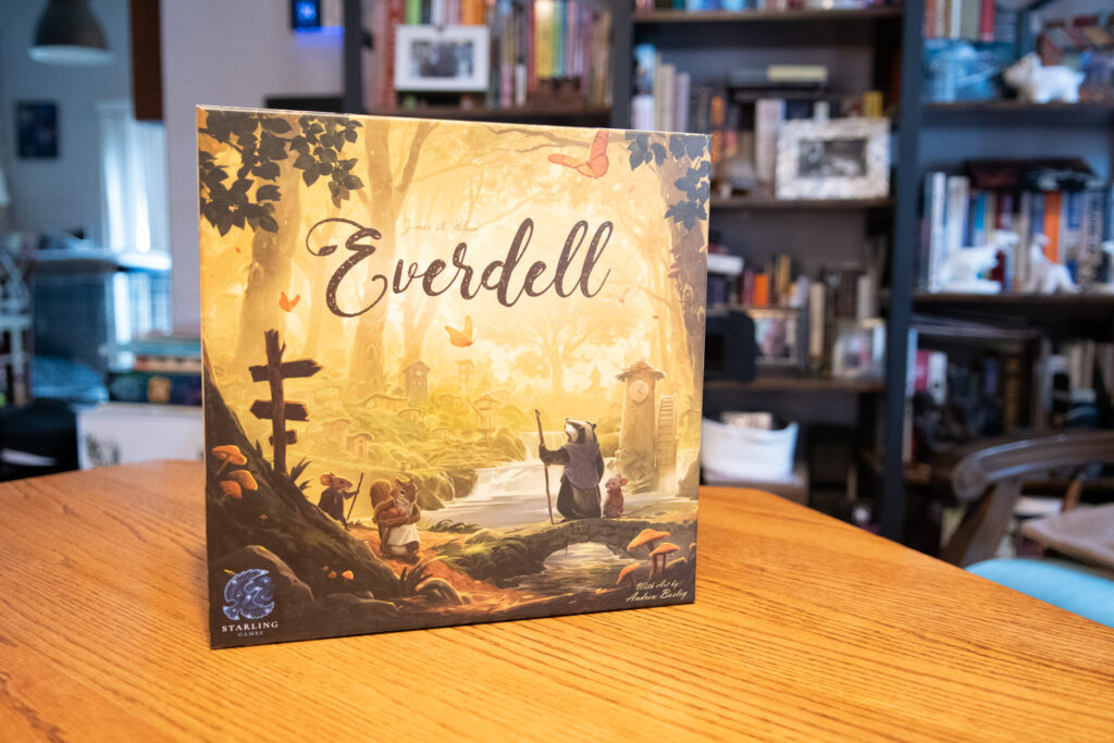 The game box for Everdell featuring a forest scene is on a wood table. A bookcase is in the background.