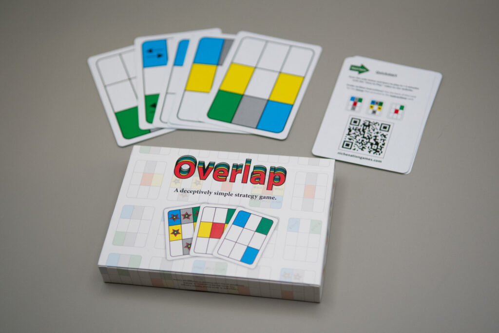 A card box reads Overlap "A deceptively simple strategy game." Three cards showing a grid on 9 boxes are under the words. Some of the boxes are colored in and some are blank. Above the box, several game cards and the Quick Start Guide for the game are out of the box.