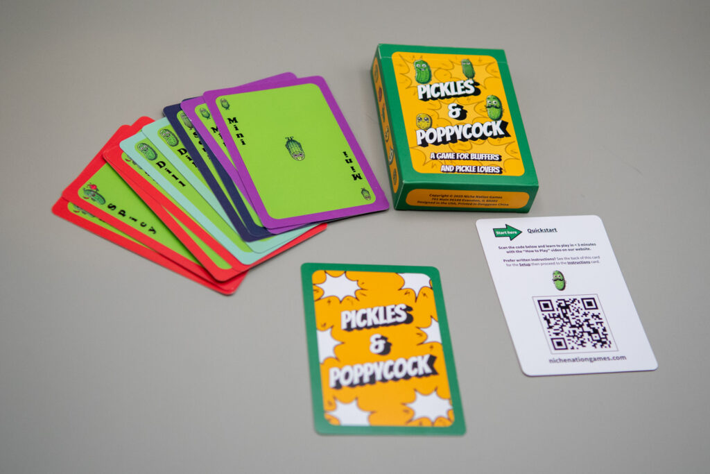 A card box reads "Pickles & Poppycock A Game for Bluffers and Pickle Lovers." Also shown are several face up game cards picturing different types of pickles, a card that reads Pickles & Poppycock and a card that has a quick start guide and QR code.