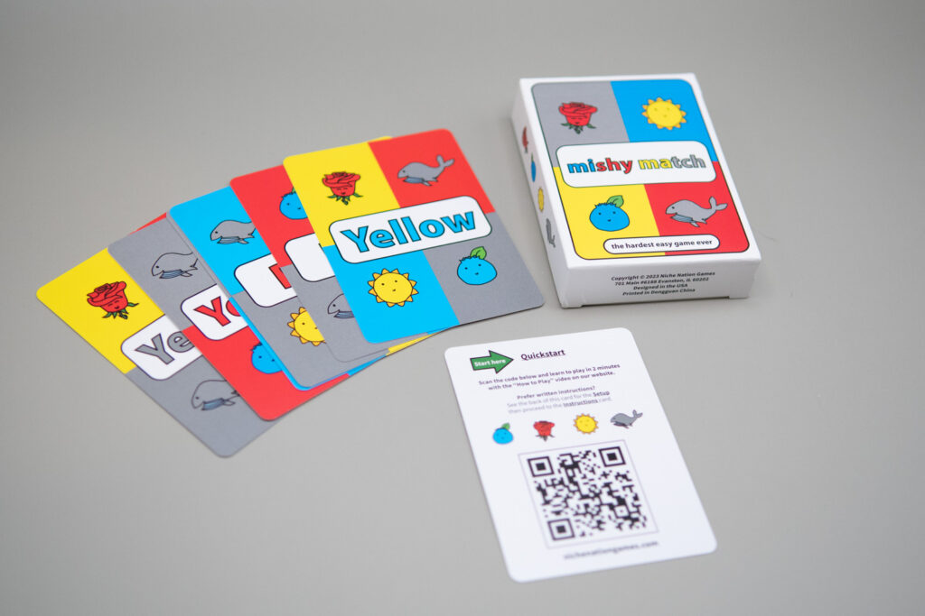 A card box reads the words mishy match "the hardest easy game ever." Pictured on the box are a red rose, a yellow sun, a blue berry and a gray whale. Spread out are several of the playing cards. In the center of the cards is a color word spelled out.